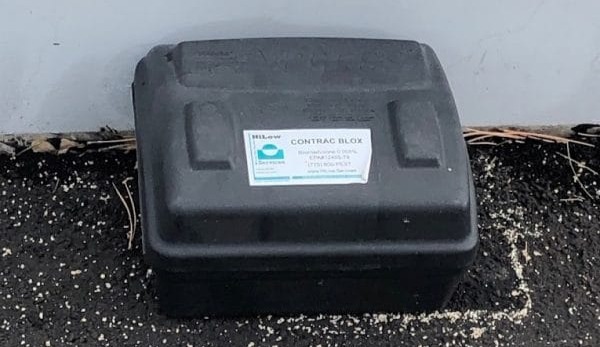 Black plastic utility box with a white label, mounted on a paved surface against a light wall.